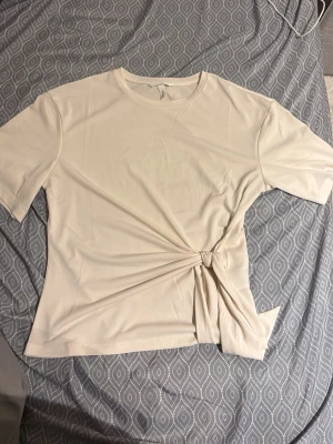 Beige topp med knyt i sidan - Säljer en stilren beige topp med rund halsringning och korta ärmar. Toppen har en snygg knytdetalj i sidan som ger en unik twist till outfiten. Perfekt att matcha med både jeans och kjol för en trendig look.