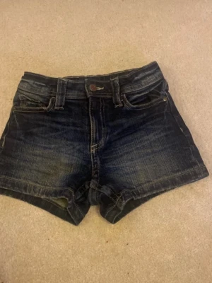 Mörkblå jeansshorts med fickor - Snygga mörkblå jeansshorts i klassisk femficksmodell med slitningar och knappgylf. Shortsen har två bakfickor med lock och knappdetaljer samt bälteshällor. Perfekta till sommaren och lätta att matcha med olika toppar.