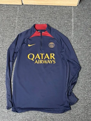 Mörkblå PSG träningströja Nike - Snygg mörkblå långärmad träningströja från PSG med gul Nike-logga och Qatar Airways-tryck på bröstet. Tröjan har halv dragkedja, röd detalj vid kragen och diskreta mönster på ärmarna. Perfekt för fotbollsträning eller chill.