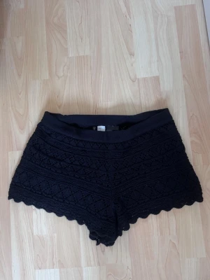 Svarta virkade shorts med spetskant - Snygga svarta shorts med virkad struktur och dekorativ spetskant nedtill. De har en elastisk midja och är perfekta för varma dagar. Passar dig som gillar en feminin och avslappnad stil.