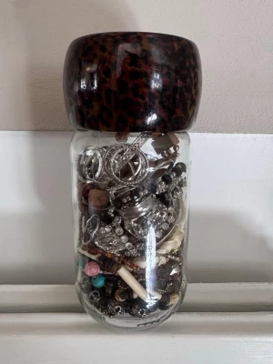 Blandade smycken i glasburk - En glasburk fylld med olika smycken som armband, halsband och ringar i blandade färger och material. Innehåller både metall, plast och textil. Perfekt för dig som vill ha unika accessoarer till din stil.