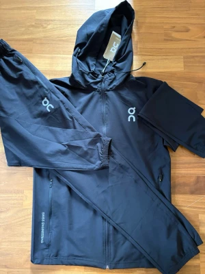 on cloud tracksuit - Snyggt svart träningsset från On med matchande jacka och byxor. Jackan har huva, dragkedja och två fickor med dragkedja. Byxorna har resår i midjan och vid benslut samt logga på benet. Materialet är lätt och funktionellt, perfekt för träning.