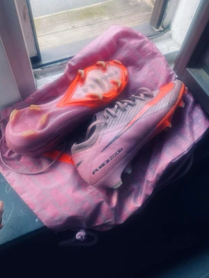 Nike Air Zoom rosa fotbollsskor - Säljer ett par Nike Air Zoom fotbollsskor i rosa med orange detaljer och svarta loggor. Skorna har snörning, låg profil och dobbar för spel på gräs. Tillverkade i syntetmaterial för lätt känsla och bra grepp på planen. Perfekta för dig som vill sticka ut på matchen!