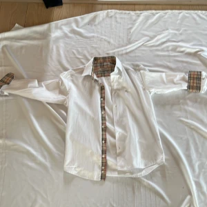 Vit skjorta med rutiga detaljer från Burberry - Säljer en klassisk vit skjorta från Burberry med ikoniska beige-rutiga detaljer på insidan av kragen, knappslån och ärmsluten. Skjortan har lång ärm, knappar framtill och en liten broderad logga på bröstet. Perfekt för dig som gillar stilrena plagg med lyxig känsla.