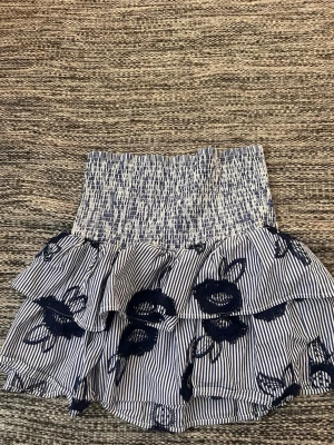 Blåvit volangkjol Holly & Whyte - Blåvit kortkjol från Holly & Whyte med smock i midjan, volanger och tunna ränder. Kjolen har broderade blommor i mörkblått. Ordinarie pris, ca 300 kr. Knappt använd!