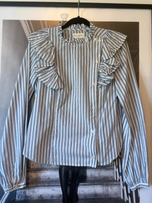 Blårandig blus med volanger Holly & White - Snygg blus från Holly & White med blå och vita vertikala ränder. Blusen har långa ärmar, volangdetaljer vid axlar och bröst samt knäppning framtill. 