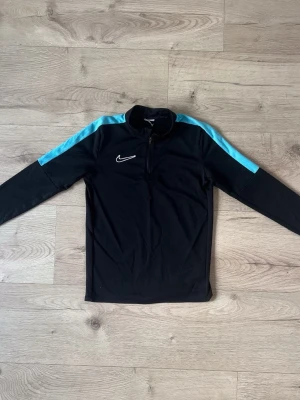 Svart Nike träningströja med blå detaljer - Snygg svart långärmad träningströja från Nike med ljusblå ränder längs axlarna och dragkedja vid halsen. Tillverkad i ett lätt och funktionellt material som passar perfekt till träning eller uppvärmning. Nike-logga broderad på bröstet.