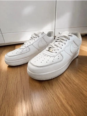 Vita Nike Air Force 1 sneakers - Klassiska vita Nike Air Force 1 sneakers med lågt skaft och ikonisk swoosh på sidan. Skorna har perforerad tå för ventilation, vita skosnören och en tjock sula. Tillverkade i läder med detaljer i gummi och textil på insidan.