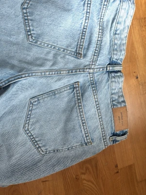 Ljusblå mom jeans med hög midja - Säljer ett par ljusblå mom jeans med hög midja och raka ben. Jeansen har klassiska fem fickor och bälteshällor. Tillverkade i kraftigt denimtyg som ger en vintagekänsla. Perfekta för en avslappnad och trendig look.