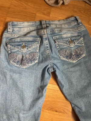 Lowwaist Ljusblå jeans med broderade fickor - Säljer ett par ljusblå jeans med snygga broderade detaljer på bakfickorna och dekorativa knappar. Jeansen har normal midja, raka ben och klassisk femficksmodell. Perfekta för dig som vill ha något unikt i garderoben!