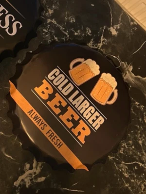 Väggdekoration öl kapsylformad - Säljer en svart och guldfärgad väggdekoration i form av en stor kapsyl med texten 'Cold Larger Beer Always Fresh' och två ölglas. Tillverkad i metall och har en upphängningsögla på baksidan. Perfekt för att ge köket eller hemmabaren en cool vibe.