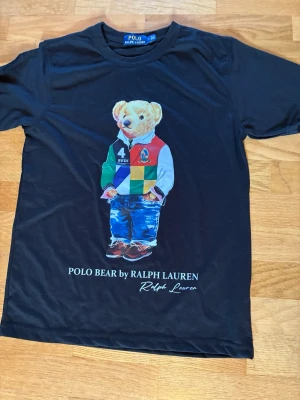 Svart Polo Bear t-shirt från Ralph Lauren - Svart t-shirt från Ralph Lauren med ikoniskt Polo Bear-tryck framtill. Björnen har färgglada kläder och texten 'POLO BEAR by RALPH LAUREN' under motivet. Klassisk rund hals och korta ärmar. Perfekt för dig som gillar streetstyle och vill sticka ut.       Kontakta gärna vid frågor och prisförslag. Skickar inom 24h