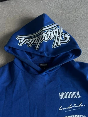 Blå tracksuit från Hoodrich med tryck - Snygg blå hoodie från Hoodrich med flera vita tryck på bröstet och ärmarna samt stor logga på huvan. Klassisk känguruficka framtill och ribbade muddar. Perfekt för dig som gillar streetwear och vill sticka ut med coola detaljer.