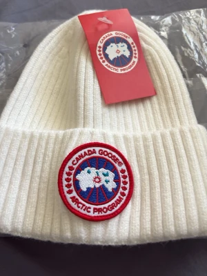 Vit ribbstickad mössa Canada Goose - Snygg vit ribbstickad mössa från Canada Goose med klassisk rund patch framtill i rött, blått och vitt. Mössan har uppvikt kant och är tillverkad i ett mjukt, värmande material som passar perfekt för kalla dagar.