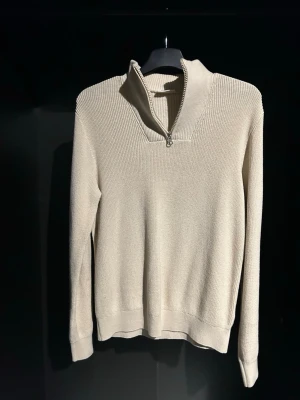 Beige stickad tröja med halv dragkedja - Snygg beige stickad tröja med halv dragkedja och hög krage. Tröjan har lång ärm och ribbade muddar vid ärmslut och nederkant. Perfekt för lager på lager och enkel att matcha med olika outfits.   Strlk 146/152