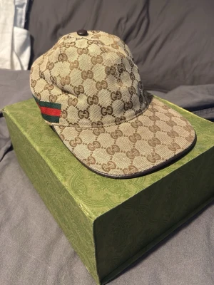 Beige GG keps från Gucci - Säljer en beige keps från Gucci med klassiskt GG-monogram i brunt över hela kepsen. På sidan finns en grön och röd rand som signaturdetalj. Kepsen har böjd skärm och är tillverkad i canvasmaterial. Perfekt för dig som vill ha en lyxig och trendig accessoar.