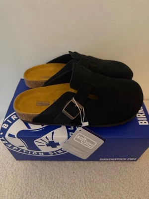 Svarta Birkenstock Boston tofflor 37 - Svarta Birkenstock Boston tofflor i mocka med klassisk design och justerbart spänne i metall. Fotbädden är i kork och insidan har en mjuk gulbrun yta. Perfekta för en chill och stilren look hemma eller ute.