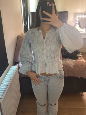Ljusblå blus med volang och knappar - Supersöt ljusblå blus med volangkant och knappar framtill. Blusen har långa puffärmar och smockdetaljer över hela bröstet och midjan. Perfekt för dig som gillar en feminin och trendig stil. Använt 2 gånger:)