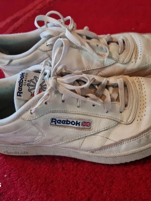 Vita Reebok Classic sneakers - Klassiska vita Reebok sneakers i läder med lågt skaft och ikonisk logga på sidan. Skorna har vita snören, perforerad tå och diskret Union Jack-detalj. Perfekta för dig som gillar retrostil och tidlös design.