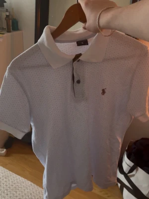 Vit pikétröja från Polo Sport Ralph Lauren - Klassisk vit pikétröja från Polo Sport Ralph Lauren med korta ärmar och diskret prickigt mönster. Tröjan har krage, knappar framtill och den ikoniska loggan broderad på bröstet. Perfekt till jeans eller shorts för en clean look.