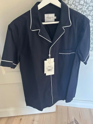 Mörkblå kortärmad skjorta Les Deux - Stilren mörkblå skjorta från Les Deux med korta ärmar och vita kontrastsömmar längs krage, knappslå och bröstficka. Klassisk look med avslappnad passform och knappar framtill. Helt oanvänd och nypris 899kr. 