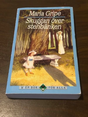 Skuggan över stenbänken av Maria Gripe - En klassisk roman av Maria Gripe med blått mjukt omslag. Boken har ett illustrerat motiv på framsidan med två kvinnor i gammaldags kläder vid en stenbänk i en skog. Utgiven av En bok för alla. Perfekt för dig som gillar mystik och spänning.