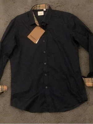 Mörkblå skjorta från Burberry - Snygg mörkblå skjorta från Burberry med klassiskt rutigt mönster på insidan av kragen och manschetterna. Skjortan har knappar framtill och en diskret broderad logga på bröstet. Perfekt för dig som gillar stilrena detaljer och exklusiv känsla. Med skjortan följs det med en enkel påse med burberry som titel.