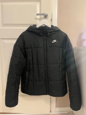 Svart pufferjacka från Nike med huva - Säljer en snygg svart pufferjacka från Nike med huva och dragkedja framtill. Jackan har en stilren design med Nike-logga på bröstet och quiltad struktur. Varm och skön att bära, perfekt för kalla vinterdagar. Lite fläckar på luvan (syns på sista bilden)