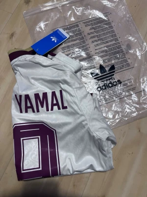 Vit Spanien fotbollströja Adidas - Säljer en vit Spanien fotbollströja från Adidas med vinröda detaljer och nummer 19 på framsidan samt Yamal och nummer 19 på ryggen. Tröjan har korta ärmar, guldfärgade muddar och officiella klubbmärken. Tillverkad i ett lätt och ventilerande material.