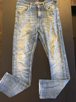 Ljusblå slitna jeans med raka ben - Säljer ett par ljusblå jeans med slitningar och hål på framsidan. De har storlek W28 och L32. Jeansen har tight passform, och klassisk femficksmodell och dragkedetaljer vid benslut. Perfekta för en stilig och trendig look.