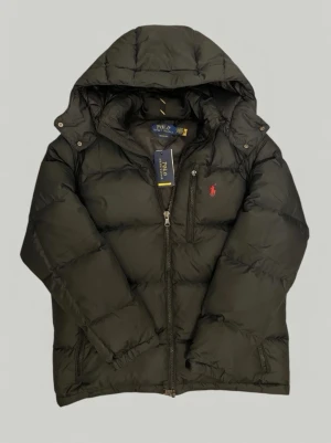 Polo Ralph Lauren Gorham - Snygg svart pufferjacka från Polo Ralph Lauren med huva och dragkedja framtill. Jackan har två sidofickor med dragkedja och den klassiska röda loggan broderad på bröstet. Perfekt för kalla vinterdagar och riktigt skön att bära.