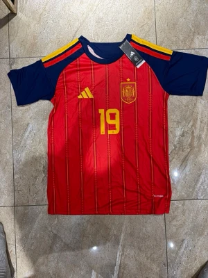 Spaniens landslagströja nr 19 - Säljer en röd och marinblå Spanien fotbollströja från Adidas med gula detaljer och nummer 19. Tröjan har LAMINE YAMAL tryckt på ryggen och officiellt landslagsemblem på bröstet. Tillverkad i lätt funktionsmaterial, perfekt för match eller träning.