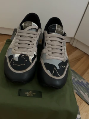 Valentino rockrunners  i svart och grått - Snygga rockrunners från Valentino med svart, grå och vit camo-design. Skorna har beige snörning, svarta detaljer och en robust sula. Materialmix av mocka och textil ger en exklusiv känsla. Perfekta för dig som vill sticka ut med stilrena detaljer.