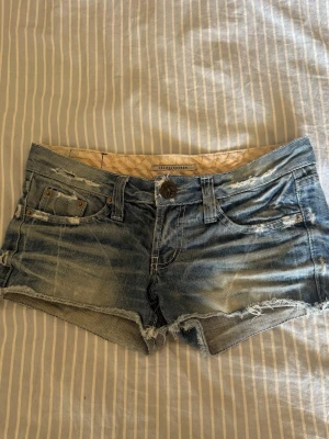 Blå jeansshorts med råa kanter - Säljer ett par blå jeansshorts med slitna detaljer och råa kanter. Shortsen har fem fickor, bälteshällor och knappgylf. Perfekta för varma dagar och ger en avslappnad vibe.
