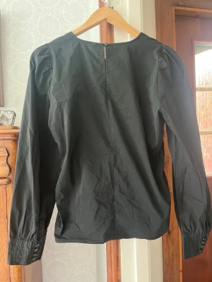 Svart blus med rynk framtill - Stilren svart blus med rund halsringning och rynkdetalj framtill. Blusen har långa ärmar med breda manschetter och knappar vid ärmsluten. Materialet är tunt och lätt, perfekt för en trendig look.