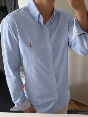 Ljusblå skjorta från Ralph Lauren - Klassisk ljusblå skjorta från Ralph Lauren med button-down krage och broderad logga på bröstet. Skjortan har långa ärmar, vita knappar och randigt tyg på insidan av manschetterna. Perfekt för en clean och stilren look. Storlek m men sitter som S 