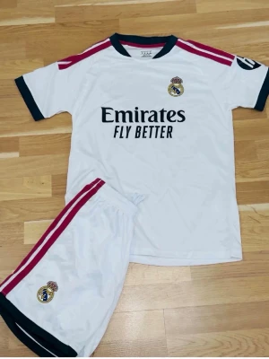 Real Madrid matchställ i vitt - Säljer ett vitt Real Madrid matchställ med kortärmad tröja och shorts. Tröjan har svarta detaljer vid hals och ärmslut, röda ränder på axlarna och klubbmärke på bröstet. Shortsen har röda ränder på sidan och klubbmärke. Perfekt för fotbollsträning eller match.