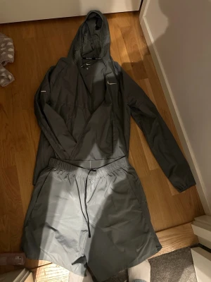 Grå träningsset från Nike - Säljer ett grått träningsset från Nike med en lättviktig vindjacka med huva och dragkedja samt matchande shorts med dragsko i midjan. Båda plaggen har en sportig design och är tillverkade i ett tunt, snabbtorkande material. Perfekt för löpning eller gym.
