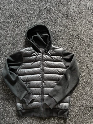 Svart cardigan från Polo Ralph Lauren - Snygg svart cardigan från Polo Ralph Lauren med huva och dragkedja. Jackan har quiltad framsida i glansigt material och ärmar samt huva i mjukare tyg. Perfekt för kalla vinterdagar och har en stilren look.