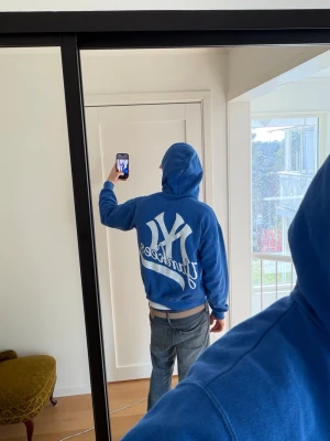 Blå Yankees hoodie med tryck - Säljer en blå hoodie med stort vitt yankees tryck på ryggen. Klassisk huva och känguruficka framtill. Perfekt för dig som gillar sportig och avslappnad stil.