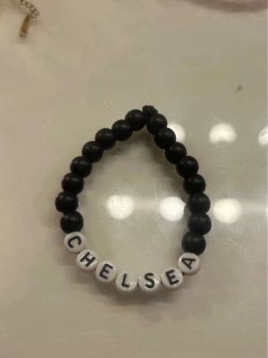Svart pärlarmband med text CHELSEA - Säljer ett svart pärlarmband med vita bokstavspärlor som stavar CHELSEA. Armbandet är elastiskt och består av runda svarta pärlor samt vita pärlor med svarta bokstäver. Perfekt för dig som vill ha ett personligt smycke.