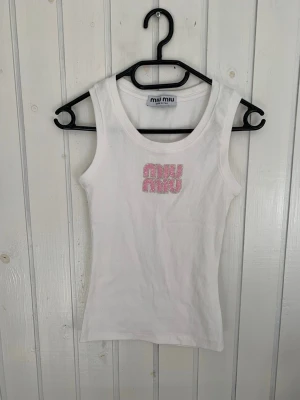 Vit ribbad linnetopp från Miu Miu - Säljer en vit ribbad linnetopp från Miu Miu med glittrande rosa logga framtill. Toppen är ärmlös och har en tight passform, perfekt för en trendig och enkel look. Materialet känns mjukt och stretchigt.