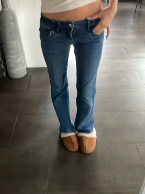 Blå bootcut jeans med låg midja - Säljer ett par blå bootcut jeans med låg midja och klassiska fem fickor. Jeansen har en lätt tvättad look och subtila slitningar vid fickorna. Perfekta för dig som gillar en avslappnad och trendig stil. Säljer för att de är för små.