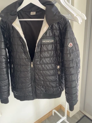 Svart dunjacka från Moncler - Snygg svart dunjacka från Moncler med quiltat mönster, hög krage och dragkedja framtill. Jackan har två sidofickor med dragkedja och en Moncler-logga på bröstet samt ärmen. Perfekt för kalla vinterdagar och riktigt stilren.
