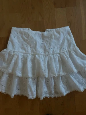 Vit volangkjol med spetsdetaljer - Söt vit kortkjol med dubbla volanger och broderade spetsdetaljer. Kjolen har ett blommigt hålmönster och hög midja, perfekt för en somrig och romantisk look. 