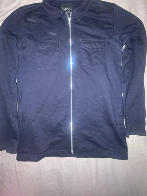 Mörkblå overshirt från boohooMAN - Stilren mörkblå overshirt från boohooMAN med dragkedja framtill och två bröstfickor med lock. Skjortkrage och långa ärmar. Tillverkad i mjukt bomullsliknande material som känns bekvämt och passar perfekt till lager-på-lager.
