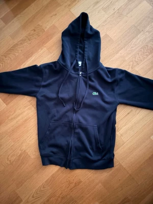  Lacoste - Säljer en svart hoodie från Lacoste med dragkedja framtill och klassisk huva med snören. Tröjan har två fickor på magen och den ikoniska gröna krokodilloggan broderad på bröstet. Perfekt för en chill och stilren look.