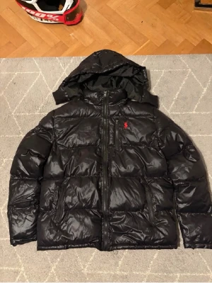 Svart dunjacka med huva från Polo Ralph Lauren - Säljer en svart dunjacka från Polo Ralph Lauren med blank finish och röd logga på bröstet. Jackan har huva, dragkedja framtill och flera praktiska fickor. Perfekt för kalla vinterdagar och riktigt snygg streetstil.