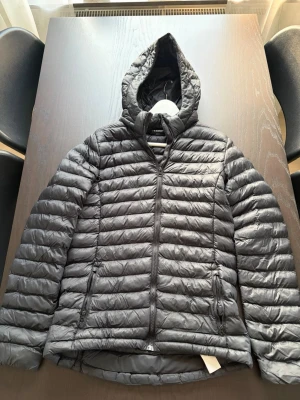 Svart dunjacka från Everest - Säljer en svart dunjacka från Everest med huva och dragkedja framtill. Jackan har två sidofickor med dragkedja och quiltad design som ger en sportig look. Perfekt för kalla vinterdagar och riktigt skön att bära.