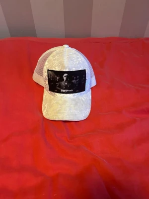 Vit meshkeps med svart tryck - Säljer en vit keps med mesh baktill och ett svart tryck framtill som föreställer en man i kostym. Kepsen har böjd skärm och justerbar storlek bak. Perfekt för dig som gillar streetwear och vill sticka ut med en unik detalj.